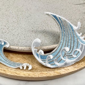 May include: Two embroidered iron-on patches depicting stylised blue waves with white outlines. The waves are in different sizes and shapes.