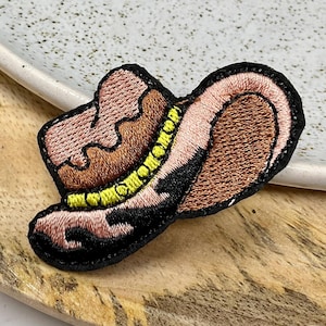 Peut inclure: Un chapeau de cowboy brodé en marron et rose avec un bandeau jaune. Le chapeau est sur une surface en bois.