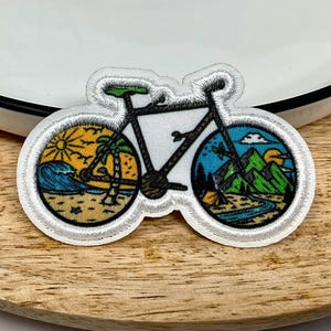 Peut inclure: Écusson brodé en forme de vélo. Les roues présentent des paysages colorés : une plage avec un palmier et une chaîne de montagnes avec une rivière. Le cadre du vélo est noir avec une selle verte.