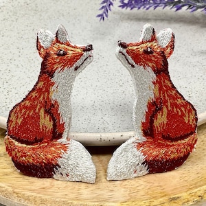 May include: Two embroidered fox patches, one facing left and one facing right, are sitting on a wooden plate. The foxes are red and orange with white accents.
