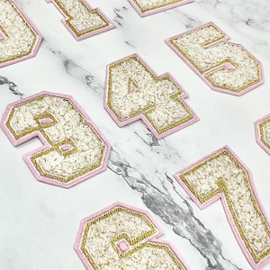 Peut inclure: Ensemble de patchs numérotés brodés, chacun avec un remplissage texturé blanc, une bordure dorée et un contour rose. Les chiffres de 0 à 9 sont présentés, parfaits pour la personnalisation de vêtements.