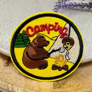 Peut inclure: Un écusson brodé jaune et noir avec un ours brun, une personne, un feu de camp et une tente. Le texte "Camping" est en lettres rouges.