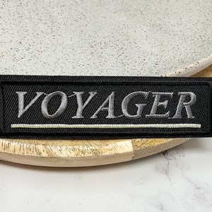 Peut inclure: Écusson en tissu noir avec des coutures blanches. L'écusson a le mot "VOYAGER" brodé en fil argenté.