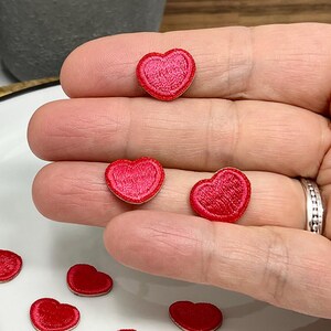 Peut inclure: Petites pièces en forme de cœur rouge brodé. Plusieurs pièces sont éparpillées sur une assiette blanche, tandis que d'autres sont tenues dans une main. Les cœurs ont un contour cousu et un remplissage texturé, idéal pour l'artisanat.