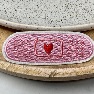 Peut inclure: Patch en tissu brodé rose en forme de pansement avec un cœur rouge au centre.