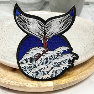 May include: An embroidered patch depicting a whale tail emerging from blue water with white waves. The patch is white and blue and is on a wooden surface.