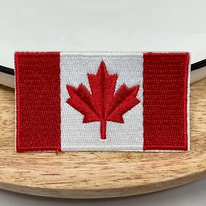 May include: Embroidered patch depicting the Canadian flag. The rectangular patch has red vertical stripes on each side and a white centre with a red maple leaf. The patch is placed on a wooden surface.