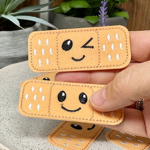 May include: Three embroidered felt patches shaped like bandages with different facial expressions. The top bandage has a winking eye, the middle bandage has a smiling face, and the bottom bandage has a closed eye.