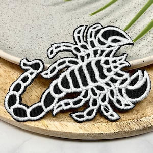 May include: Black and white embroidered scorpion patch. The scorpion is outlined in white and filled in with black thread. The patch is on a wooden surface.