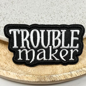 May include: Black embroidered patch with white lettering that reads "Trouble maker".
