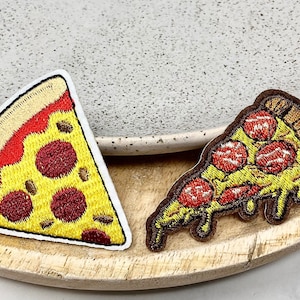 May include: Two embroidered pizza slice patches. One is yellow with red pepperoni and a white crust. The other is brown with red pepperoni and a white crust.