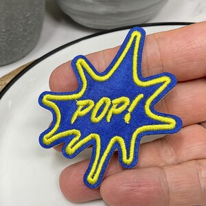 May include: An iron-on patch embroidered in blue and yellow with the word "POP!" in a yellow outline.