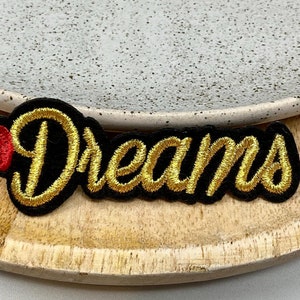 May include: Gold embroidered "Dreams" patch with a red heart. The patch is on a wooden surface.