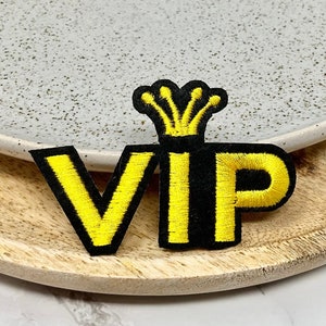 May include: Yellow embroidered VIP patch with a black crown on top. The patch is on a wooden surface.
