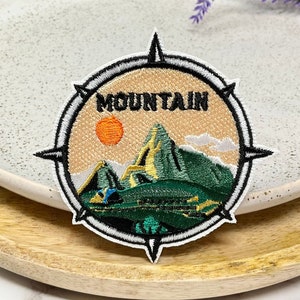Peut inclure: Un écusson brodé en forme de boussole blanc et noir avec une scène de montagne verte, jaune et orange. Le texte "MOUNTAIN" est brodé en noir sur l'écusson.
