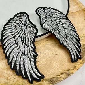 May include: Two silver embroidered angel wings with black fabric backing. The wings are on a wooden surface.