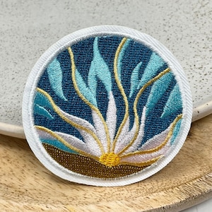 May include: Circular embroidered patch with a floral design. The patch has a dark blue background with light blue, white, and gold embroidery. It is edged with a white border. The patch is likely for clothing.