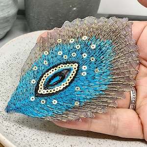 Könnte beinhalten: Ein blauer und goldener Pfauenaugen-Stickerei-Patch mit Pailletten. Der Patch befindet sich auf einem transparenten Hintergrund und wird von einer Hand gehalten.