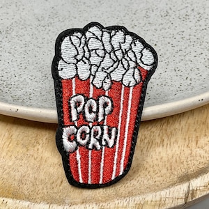 May include: A red and white embroidered iron-on patch with the words "POP CORN" in white and a white embroidered image of popcorn in a red and white striped container.