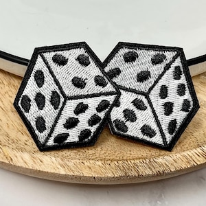 May include: Two embroidered dice patches with a white background and black dots, outlined in black. The dice are hexagonal and placed on a wooden tray, near a white plate with a black rim. These patches are suitable for clothing or accessories.