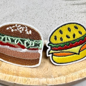 May include: Two embroidered iron-on patches of hamburgers. One is brown with green lettuce and red tomato. The other is yellow with green lettuce and red tomato.