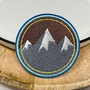 Peut inclure: Un écusson rond brodé avec une bordure bleue, jaune et verte. Le centre de l'écusson présente un fond brun avec trois montagnes blanches.