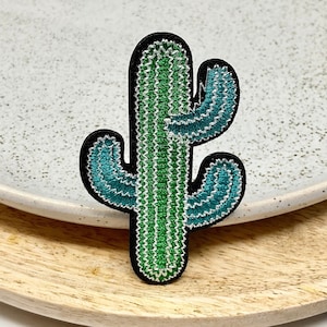May include: Embroidered cactus patch with a black border. The cactus is green with teal accents and white stitching. The patch is on a white plate and a wooden tray.