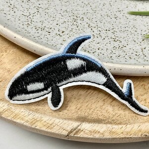 May include: Black and white embroidered orca whale patch. The patch is on a wooden surface.