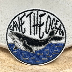 Può includere: Toppa ricamata bianca con una balena nera che nuota in acqua blu. Il testo "Save The Ocean" è scritto in nero intorno alla balena.