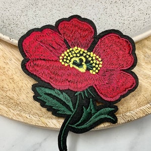 Peut inclure: Patch brodé de fleurs rouges avec des feuilles vertes et un centre jaune. Le patch est sur une surface en bois.