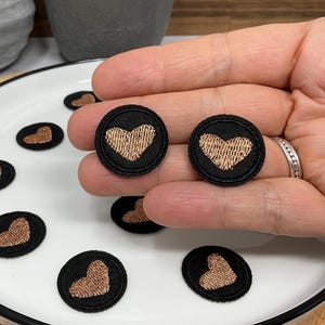 Puede incluir: Parches bordados con forma de corazón en negro y dorado. Los parches son redondos, con un corazón dorado sobre fondo negro. Varios están dispuestos sobre un plato blanco con borde negro.