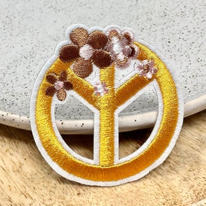 Peut inclure: Écusson brodé représentant un symbole de paix doré avec une bordure blanche, orné de fleurs marron et roses. L'écusson est posé sur une assiette grise claire mouchetée et un plateau en bois.