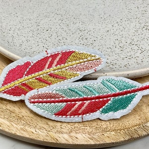 May include: Two embroidered felt feathers, one with red, yellow and pink stripes, and the other with red, green and blue stripes.