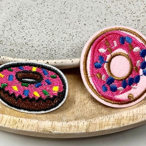 Peut inclure: Deux patchs de beignets brodés. Un beignet est rose avec des pépites bleues et blanches et un contour brun. L'autre beignet est rose avec des pépites jaunes, vertes et bleues et un contour brun.