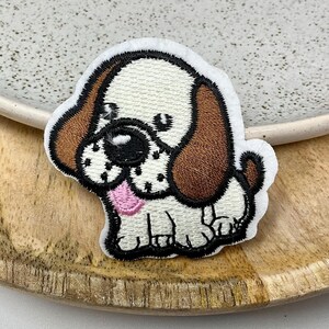 Peut inclure: Écusson brodé représentant un chiot de dessin animé avec des oreilles marron, un nez noir et une langue rose. L'écusson a un fond blanc et un contour noir. Le chiot est assis. L'écusson est sur une surface en bois.