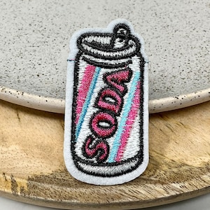 Pode incluir: Etiqueta branca bordada para passar a ferro de uma lata de refrigerante com a palavra "Soda" escrita em letras rosa e azul.