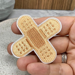 Peut inclure: Un patch thermocollant en tissu beige en forme de pansement avec des coutures blanches autour des bords.