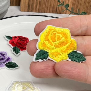 Peut inclure: Écussons de roses brodées en jaune, rouge, violet et blanc. La rose jaune est tenue dans une main, tandis que les autres sont sur une assiette blanche. Chaque écusson comporte des feuilles vertes et une bordure blanche.