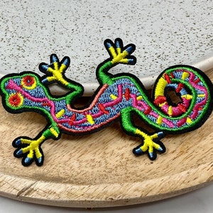 Peut inclure: Un patch brodé coloré représentant un lézard vert avec des détails jaunes, roses et bleus. Le lézard est enroulé dans une pose détendue.