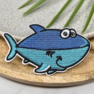 May include: Embroidered patch depicting a cartoon fish in blue and teal, with a black outline. The fish has large eyes and a smiling mouth. This patch is designed to be ironed on or sewn onto fabric.