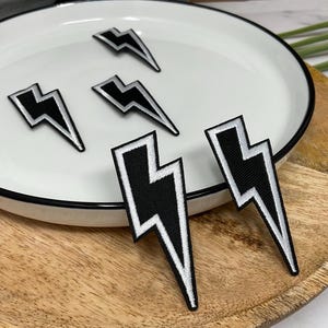 May include: Embroidered black and white lightning bolt patches. The patches are on a white plate with a black rim, set on a wooden tray. The patches have a black background with a white outline.