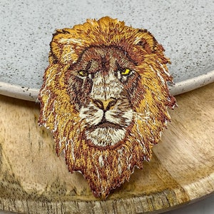 Puede incluir: Parche bordado con la cabeza de un león con un diseño detallado. El parche presenta tonos marrones y dorados, con ojos amarillos. El león está centrado en un plato gris claro y una bandeja de madera.