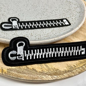 Peut inclure: Deux patchs thermocollants noirs en forme de fermetures éclair avec des coutures blanches. Les dents de la fermeture éclair sont blanches et les tirettes sont blanches.