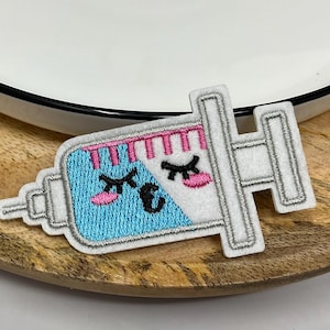 May include: An embroidered patch in the shape of a syringe with a cartoon face. The syringe is white with a blue and pink design, featuring closed eyes and a smiling mouth. The patch is placed on a wooden surface next to a white plate.