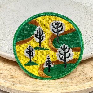 May include: A round embroidered patch with a yellow and green background. The patch features a winding brown path with five trees, four of which have white trunks and black branches. The fifth tree is a dark green pine tree.