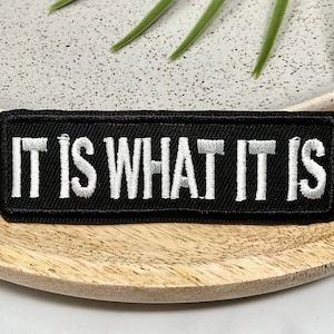 Puede incluir: Parche de tela negro con texto bordado en blanco que dice "IT IS WHAT IT IS".