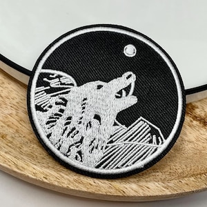 Peut inclure: Un écusson brodé noir et blanc représentant un loup hurlant à la lune. Le loup est en silhouette sur un fond noir avec des montagnes blanches et une lune blanche en haut à droite.