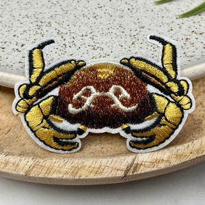 Peut inclure: Un patch brodé en forme de crabe, de couleur marron et or, avec des points de couture blancs. Le crabe est de face, les pinces levées.