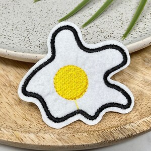 Op de afbeelding: Een geborduurde patch van wit vilt in de vorm van een spiegelei met een gele dooier.