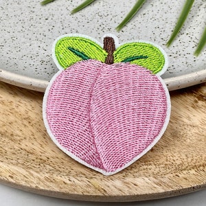 May include: A pink embroidered peach with green leaves. The peach is a popular fruit and a symbol of summer.